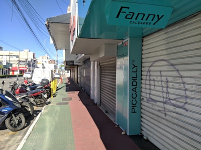 A city street view featuring several parked motorcycles and scooters near closed storefronts. The store signs include 'Fanny Calçados' and 'Piccadilly'. The sidewalk is divided into sections with green and red paving. Overhead, numerous power lines stretch across the sky.