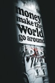 A poster with bold black text stating 'money make the world go around' dominates the upper part of the wall. Below are two smaller posters with graphics and the text 'GOOD FOR HEALTH', followed by two more posters saying 'FLY WITH ME'. The overall setting gives an urban, grunge aesthetic with dim lighting and monochromatic tones.