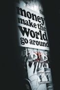 A poster with bold black text stating 'money make the world go around' dominates the upper part of the wall. Below are two smaller posters with graphics and the text 'GOOD FOR HEALTH', followed by two more posters saying 'FLY WITH ME'. The overall setting gives an urban, grunge aesthetic with dim lighting and monochromatic tones.