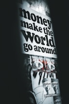 A poster with bold black text stating 'money make the world go around' dominates the upper part of the wall. Below are two smaller posters with graphics and the text 'GOOD FOR HEALTH', followed by two more posters saying 'FLY WITH ME'. The overall setting gives an urban, grunge aesthetic with dim lighting and monochromatic tones.