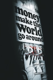 A poster with bold black text stating 'money make the world go around' dominates the upper part of the wall. Below are two smaller posters with graphics and the text 'GOOD FOR HEALTH', followed by two more posters saying 'FLY WITH ME'. The overall setting gives an urban, grunge aesthetic with dim lighting and monochromatic tones.