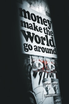 A poster with bold black text stating 'money make the world go around' dominates the upper part of the wall. Below are two smaller posters with graphics and the text 'GOOD FOR HEALTH', followed by two more posters saying 'FLY WITH ME'. The overall setting gives an urban, grunge aesthetic with dim lighting and monochromatic tones.