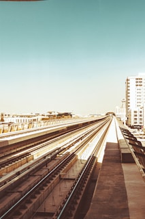 A metro railway track extends into the distance, creating a sense of perspective and depth. The tracks are flanked by buildings, including a tall white structure, under a clear sky with a gradient of blue. The urban setting is evident, and the scene is devoid of people or trains.