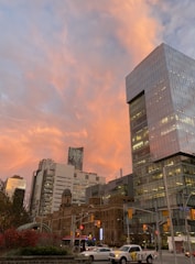A cityscape view with modern and historical buildings under a vibrant sunset sky. The skyscrapers are primarily made of glass, reflecting the orange and pink hues of the sunset. Traffic lights and several vehicles, including a bus and a pickup truck with 'INGENUITY' written on the side, are visible on the busy street below.
