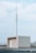 A rooftop structure with a tall metal antenna, supported by wires, rising from its top. The structure is rectangular, with a door and light fixture visible. The background consists of a pale blue sky with scattered clouds.