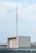A rooftop structure with a tall metal antenna, supported by wires, rising from its top. The structure is rectangular, with a door and light fixture visible. The background consists of a pale blue sky with scattered clouds.