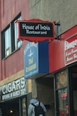 A street view of multiple restaurant signs on a building. The signs feature 'House of India Restaurant' with a red and black design, and 'Basil Court Thai Restaurant' in blue and yellow. There is also a sign indicating a cigar shop opening in March 2021, and a neon 'OPEN' sign in a window. A person with a backpack is walking towards the building.