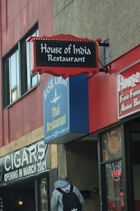 A street view of multiple restaurant signs on a building. The signs feature 'House of India Restaurant' with a red and black design, and 'Basil Court Thai Restaurant' in blue and yellow. There is also a sign indicating a cigar shop opening in March 2021, and a neon 'OPEN' sign in a window. A person with a backpack is walking towards the building.