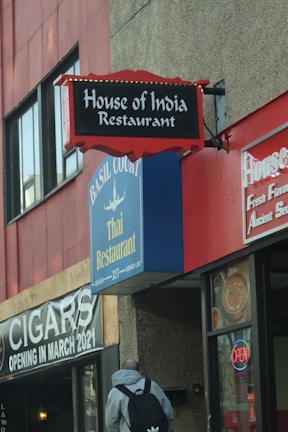 A street view of multiple restaurant signs on a building. The signs feature 'House of India Restaurant' with a red and black design, and 'Basil Court Thai Restaurant' in blue and yellow. There is also a sign indicating a cigar shop opening in March 2021, and a neon 'OPEN' sign in a window. A person with a backpack is walking towards the building.