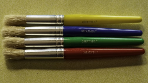 Four paintbrushes are aligned horizontally on a textured, light green surface. Each brush has a metallic ferrule and a brightly colored handle, with colors in yellow, blue, green, and red from top to bottom. The brand name 'CREATIVE PL' is printed on each handle.