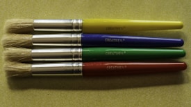 Four paintbrushes are aligned horizontally on a textured, light green surface. Each brush has a metallic ferrule and a brightly colored handle, with colors in yellow, blue, green, and red from top to bottom. The brand name 'CREATIVE PL' is printed on each handle.