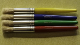 Four paintbrushes are aligned horizontally on a textured, light green surface. Each brush has a metallic ferrule and a brightly colored handle, with colors in yellow, blue, green, and red from top to bottom. The brand name 'CREATIVE PL' is printed on each handle.