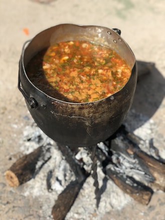 A large black pot is placed over a fire with a stew or soup simmering inside. The pot contains a colorful mixture of vegetables, herbs, and possibly meat, creating an appetizing and rustic look. Logs and ashes are visible beneath, indicating outdoor cooking.