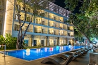 A modern, well-lit hotel building with balconies overlooks a rectangular swimming pool. The pool is illuminated by glowing, colorful spheres floating on the water's surface. Wicker lounge chairs are arranged along the poolside under lush green trees.