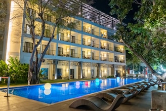 A modern, well-lit hotel building with balconies overlooks a rectangular swimming pool. The pool is illuminated by glowing, colorful spheres floating on the water's surface. Wicker lounge chairs are arranged along the poolside under lush green trees.