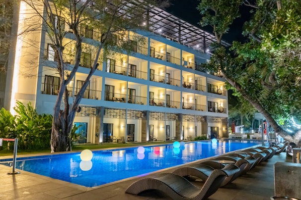 A modern, well-lit hotel building with balconies overlooks a rectangular swimming pool. The pool is illuminated by glowing, colorful spheres floating on the water's surface. Wicker lounge chairs are arranged along the poolside under lush green trees.