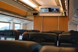 Rows of empty seats inside a train cabin, featuring modern headrests and personal screens. Overhead, a digital display board provides travel information. Large windows allow natural light to fill the space, while wooden panels add a warm touch.