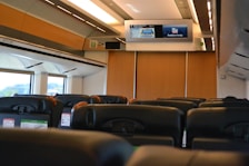 Rows of empty seats inside a train cabin, featuring modern headrests and personal screens. Overhead, a digital display board provides travel information. Large windows allow natural light to fill the space, while wooden panels add a warm touch.