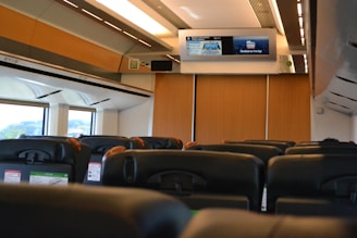 Rows of empty seats inside a train cabin, featuring modern headrests and personal screens. Overhead, a digital display board provides travel information. Large windows allow natural light to fill the space, while wooden panels add a warm touch.
