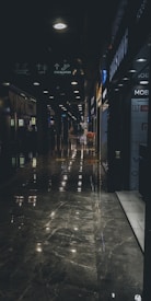 A dimly lit shopping mall corridor with reflective marble flooring. The walls are lined with various store signs, including Samsung and Oppo. The corridor appears long, with overhead lights creating a series of bright spots on the polished floor.