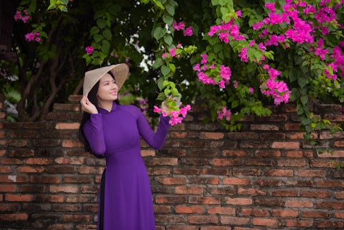 A woman wearing a traditional purple dress and a conical hat stands beside a brick wall covered with vibrant pink bougainvillea flowers. She is gently touching the flowers while looking up with a serene expression.