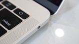 Close-up view of a laptop keyboard and speaker grille alongside a smartphone. The focus is on the laptop's 'delete' and power key, with a perforated speaker section, highlighting the sleek metallic finish. The smartphone is partially visible, emphasizing the blend of technology.