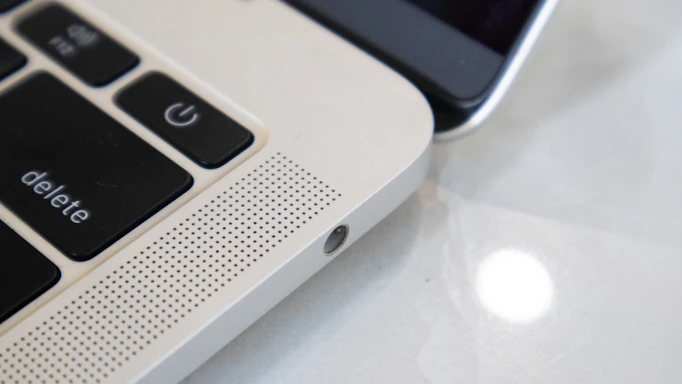 Close-up view of a laptop keyboard and speaker grille alongside a smartphone. The focus is on the laptop's 'delete' and power key, with a perforated speaker section, highlighting the sleek metallic finish. The smartphone is partially visible, emphasizing the blend of technology.
