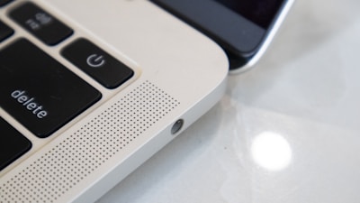 Close-up view of a laptop keyboard and speaker grille alongside a smartphone. The focus is on the laptop's 'delete' and power key, with a perforated speaker section, highlighting the sleek metallic finish. The smartphone is partially visible, emphasizing the blend of technology.