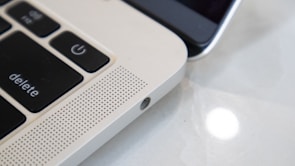 Close-up view of a laptop keyboard and speaker grille alongside a smartphone. The focus is on the laptop's 'delete' and power key, with a perforated speaker section, highlighting the sleek metallic finish. The smartphone is partially visible, emphasizing the blend of technology.