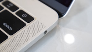 Close-up view of a laptop keyboard and speaker grille alongside a smartphone. The focus is on the laptop's 'delete' and power key, with a perforated speaker section, highlighting the sleek metallic finish. The smartphone is partially visible, emphasizing the blend of technology.