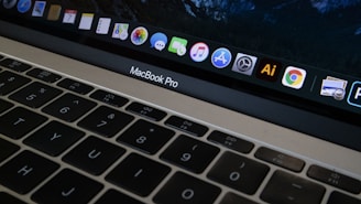A close-up view of a MacBook Pro keyboard and screen with various application icons displayed on the dock. The keyboard has a black finish with white letters, and the screen shows icons like Safari, Mail, and Finder. The lighting is dim, providing a cozy atmosphere.