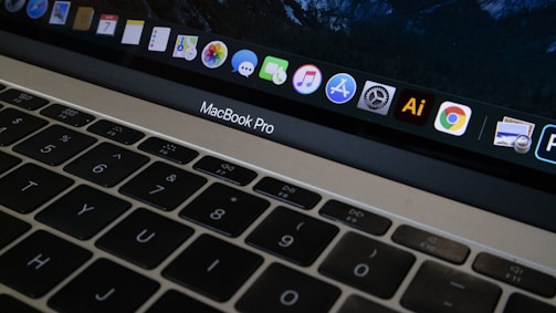 A close-up view of a MacBook Pro keyboard and screen with various application icons displayed on the dock. The keyboard has a black finish with white letters, and the screen shows icons like Safari, Mail, and Finder. The lighting is dim, providing a cozy atmosphere.