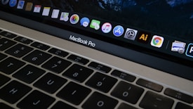 A close-up view of a MacBook Pro keyboard and screen with various application icons displayed on the dock. The keyboard has a black finish with white letters, and the screen shows icons like Safari, Mail, and Finder. The lighting is dim, providing a cozy atmosphere.