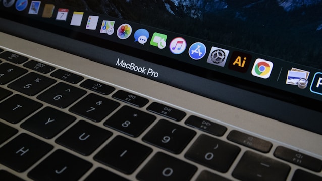 A close-up view of a MacBook Pro keyboard and screen with various application icons displayed on the dock. The keyboard has a black finish with white letters, and the screen shows icons like Safari, Mail, and Finder. The lighting is dim, providing a cozy atmosphere.