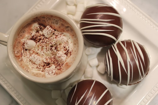 An inviting shot of a steaming cup of hot chocolate beside a plate of assorted chocolate pieces.