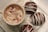 A cup of hot chocolate topped with whipped cream and sprinkled with cocoa powder, surrounded by mini marshmallows and three chocolate spheres drizzled with white chocolate, all placed on a white square plate.