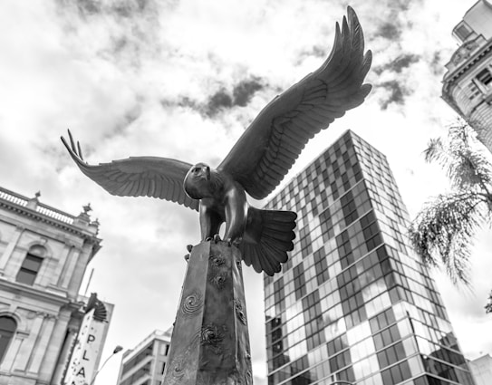 A black-and-white photograph of a large eagle statue perched atop a column, with its wings spread wide. The scene is set against a backdrop of urban architecture, including a tall modern glass building, a historical structure, and a palm tree. The sky is partly cloudy, adding depth to the image.