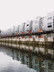 A row of modern, uniquely designed apartment buildings with a silver metallic exterior, featuring multiple small windows. The structures are built alongside a tranquil body of water reflecting them. The sky appears overcast, adding to the muted ambiance of the scene.