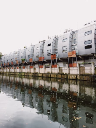 A row of modern, uniquely designed apartment buildings with a silver metallic exterior, featuring multiple small windows. The structures are built alongside a tranquil body of water reflecting them. The sky appears overcast, adding to the muted ambiance of the scene.