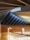 A modern architectural interior featuring a triangular wooden ceiling with exposed beams and angular lines. The walls and floor are made of light-colored wood, and a glass railing borders the walkway. Natural light streams through small windows near the ceiling, creating a serene atmosphere.