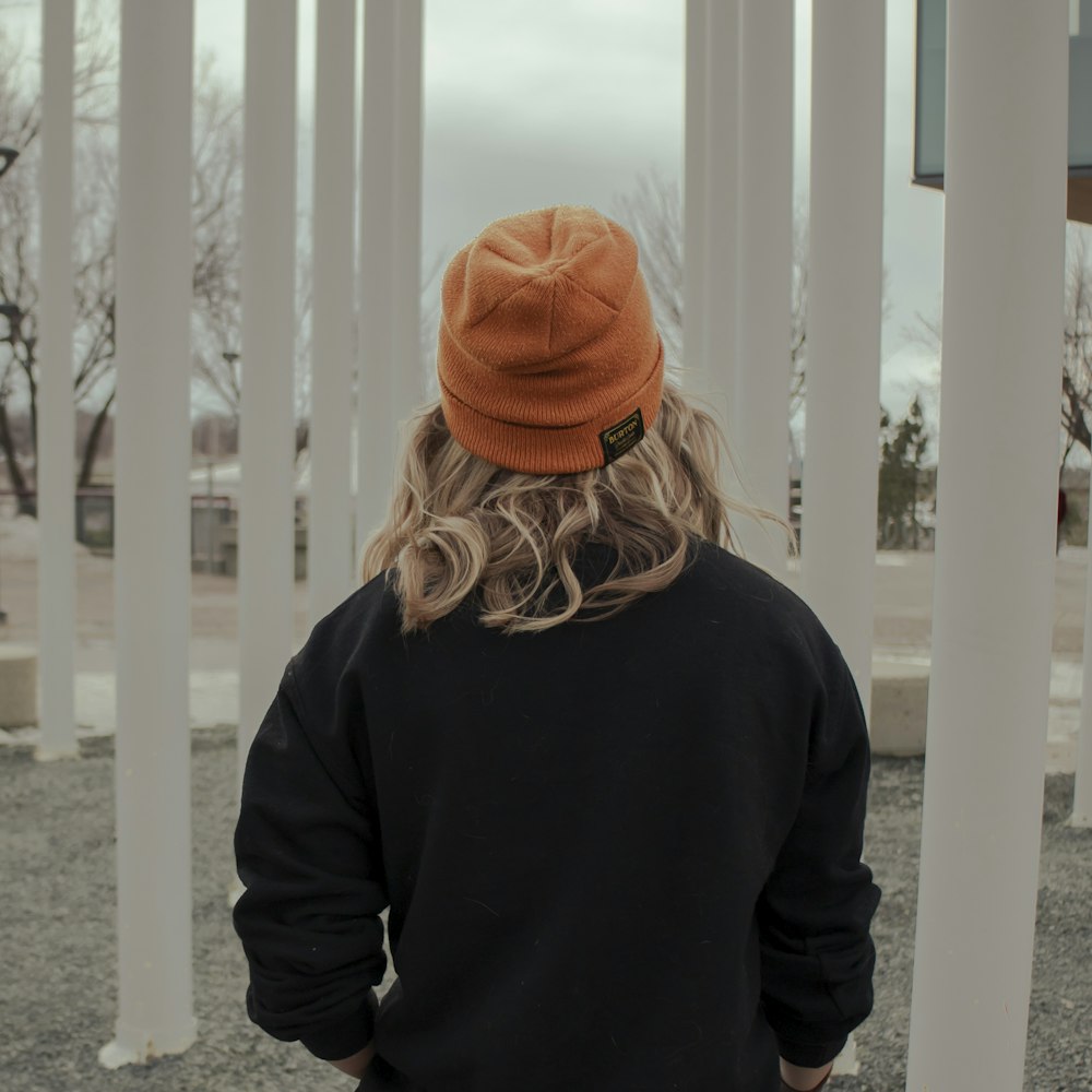 woman in black jacket and orange knit cap standing near white wooden fence during daytime