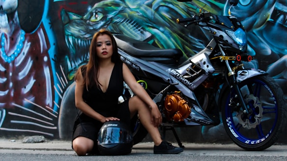 A woman with long hair is kneeling on the pavement beside a modern motorcycle. She is wearing a black dress and resting one hand on a black helmet placed on the ground. The wall behind her is covered with vibrant and intricate graffiti art featuring abstract designs and creature-like figures.