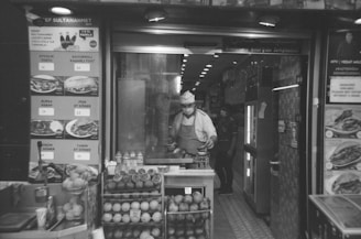 A chef wearing a mask and a uniform stands inside a small food shop or restaurant. Various food items and prices are displayed on the menu outside. Fresh fruits, possibly used for juices, are arranged near the entrance, and another person is visible in the background.