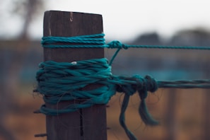 A close-up view of a dark wooden post with multiple strands of teal twine wrapped and tied around it, creating an intricate knot. The background is blurred, with subtle hues of brown and green.
