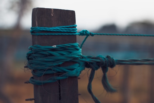 A close-up view of a dark wooden post with multiple strands of teal twine wrapped and tied around it, creating an intricate knot. The background is blurred, with subtle hues of brown and green.