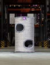 A tall, cylindrical cat tower with two openings is placed in an industrial or warehouse setting. It has a textured, grey exterior with a small purple tag bearing a logo or design.