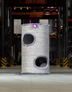 A tall, cylindrical cat tower with two openings is placed in an industrial or warehouse setting. It has a textured, grey exterior with a small purple tag bearing a logo or design.