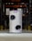 A tall, cylindrical cat tower with two openings is placed in an industrial or warehouse setting. It has a textured, grey exterior with a small purple tag bearing a logo or design.