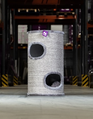 A tall, cylindrical cat tower with two openings is placed in an industrial or warehouse setting. It has a textured, grey exterior with a small purple tag bearing a logo or design.