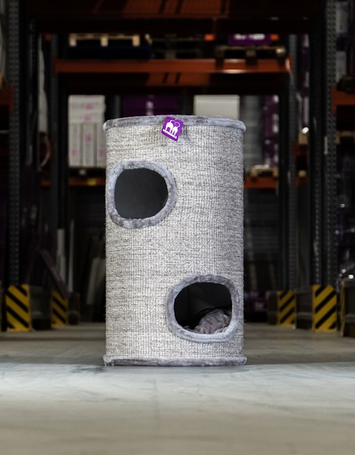 A tall, cylindrical cat tower with two openings is placed in an industrial or warehouse setting. It has a textured, grey exterior with a small purple tag bearing a logo or design.